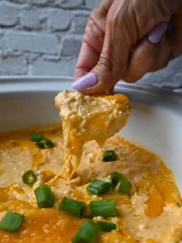 A person with lavender-painted nails dips a tortilla chip into a serving dish of cheesy ground chicken buffalo dip, which is topped with chopped green onions. The white dish sits in front of a white brick wall.