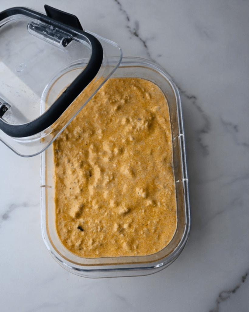 A partially open rectangular plastic container with a black-trimmed lid sits on a marble countertop, showing creamy, orange ground chicken buffalo dip inside.