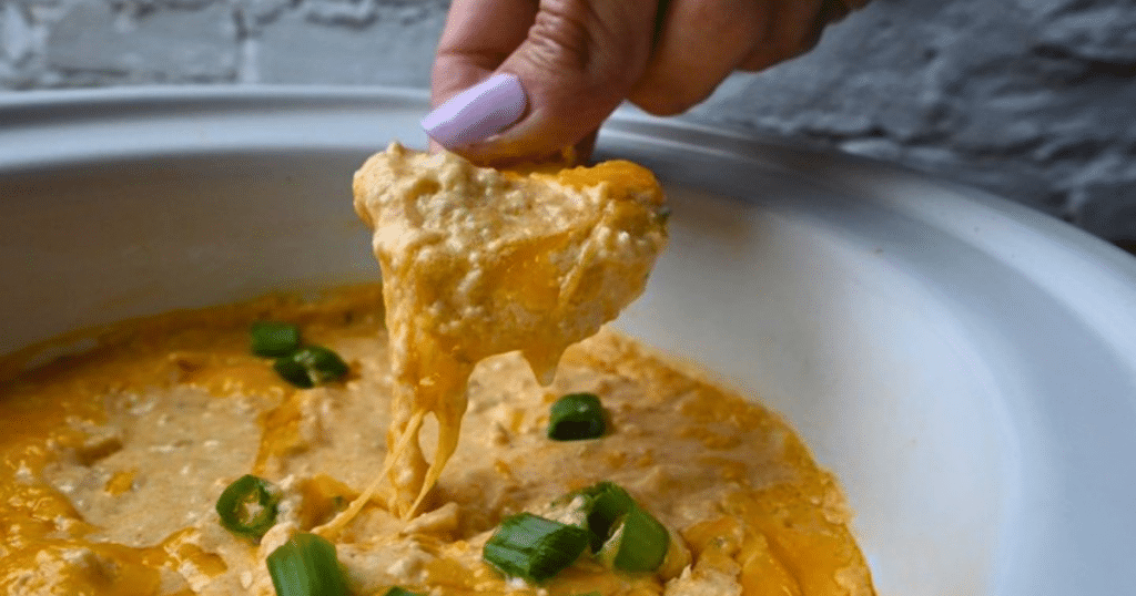 A hand with light purple polished nails dips a chip into ground chicken buffalo dip, which looks creamy and cheesy and is topped with chopped green onions, all served in a white dish.
