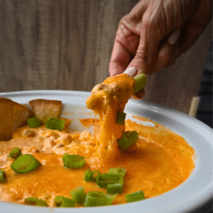 A hand dips a celery stick into Buffalo bean dip, topped with melted cheese and green onions in a white dish.