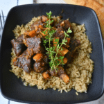 A black bowl holds seasoned rice topped with pieces of honey balsamic beef and chunks of butternut squash, finished with a sprinkle of fresh thyme. Next to the bowl are a loaf of bread and a folded napkin.