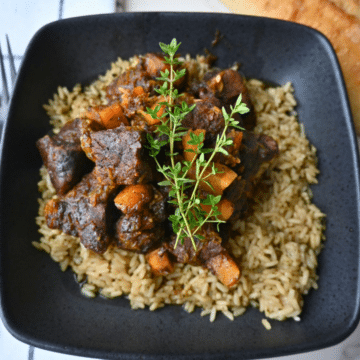 A black bowl holds seasoned rice topped with pieces of honey balsamic beef and chunks of butternut squash, finished with a sprinkle of fresh thyme. Next to the bowl are a loaf of bread and a folded napkin.