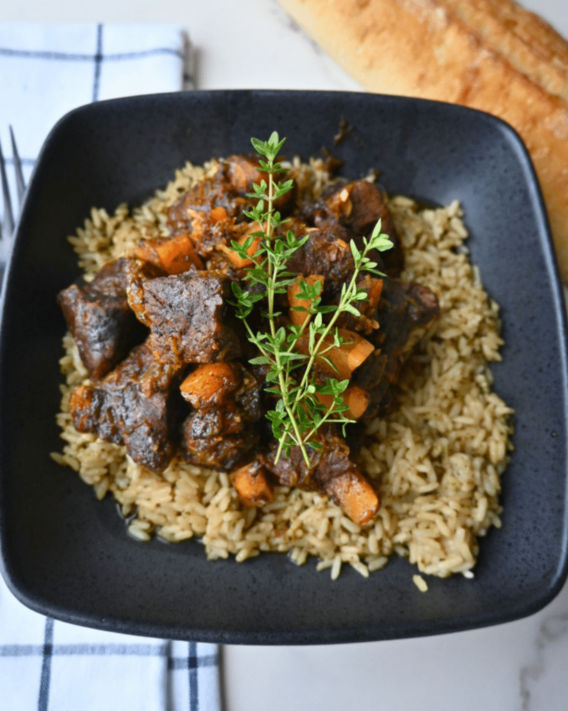 A black bowl holds seasoned rice topped with pieces of honey balsamic beef and chunks of butternut squash, finished with a sprinkle of fresh thyme. Next to the bowl are a loaf of bread and a folded napkin.