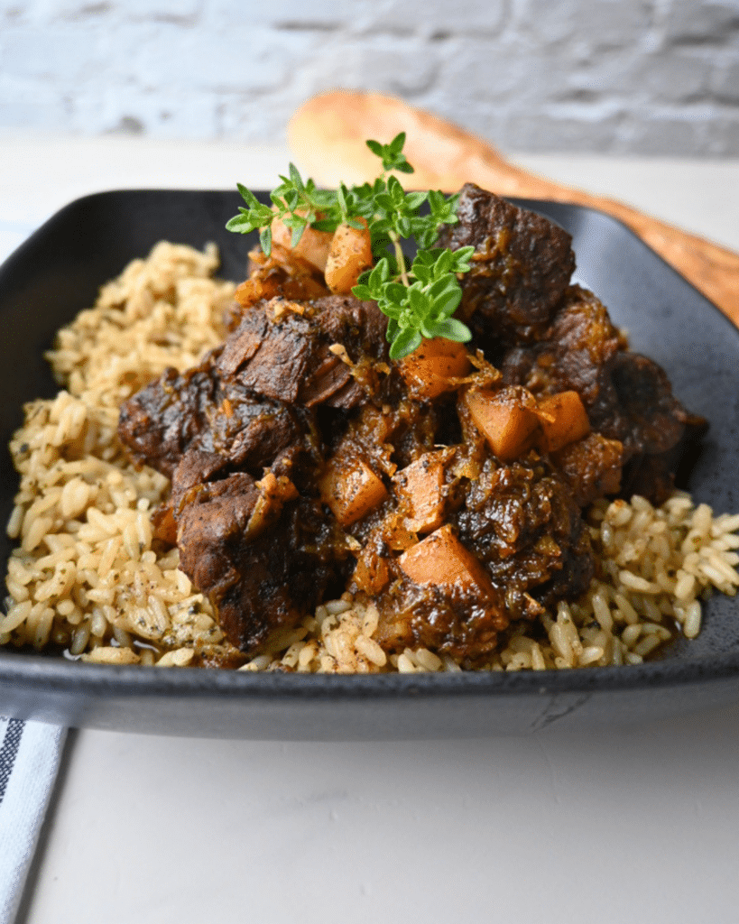 A black plate holds honey balsamic beef stew and diced butternut squash served over brown rice, topped with fresh herbs. A wooden spoon lies in the background.