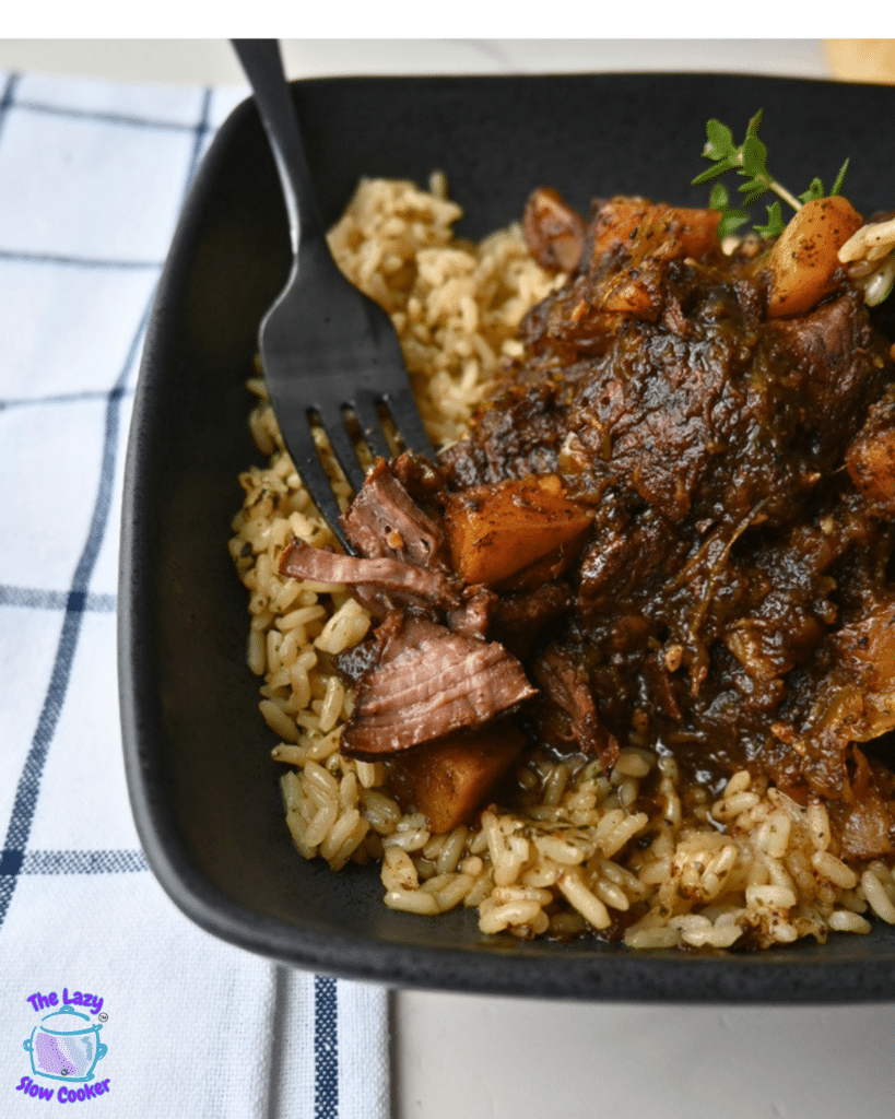 A serving of honey balsamic beef pot roast with carrots and thick gravy sits over brown rice in a black bowl, next to roasted butternut squash. A black fork rests on the bowl, which is placed on a white plaid napkin.