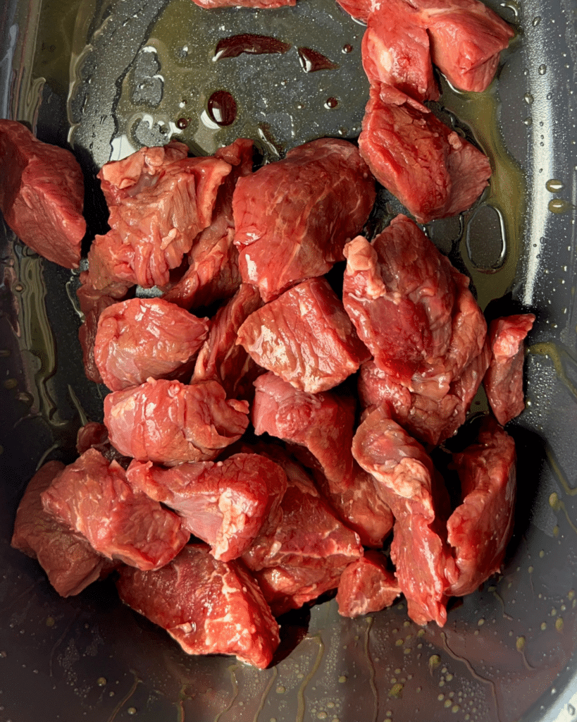 Several pieces of raw red beef sit in a dark pan lightly coated with oil, prepared for slow cooking into honey balsamic beef and butternut squash.