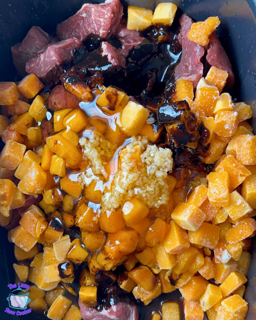 A pot holds raw beef strips, frozen diced butternut squash, minced garlic, and various sauces and seasonings all combined and ready for slow cooking into honey balsamic beef with butternut squash. In the lower left corner, there's a small logo featuring a purple chef hat.