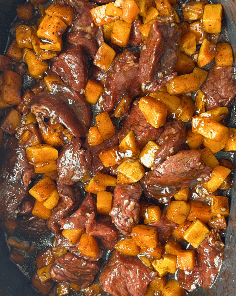 Raw beef chunks and diced butternut squash are coated in a glossy, dark marinade and mixed together in a pan, prepared for slow cooking.