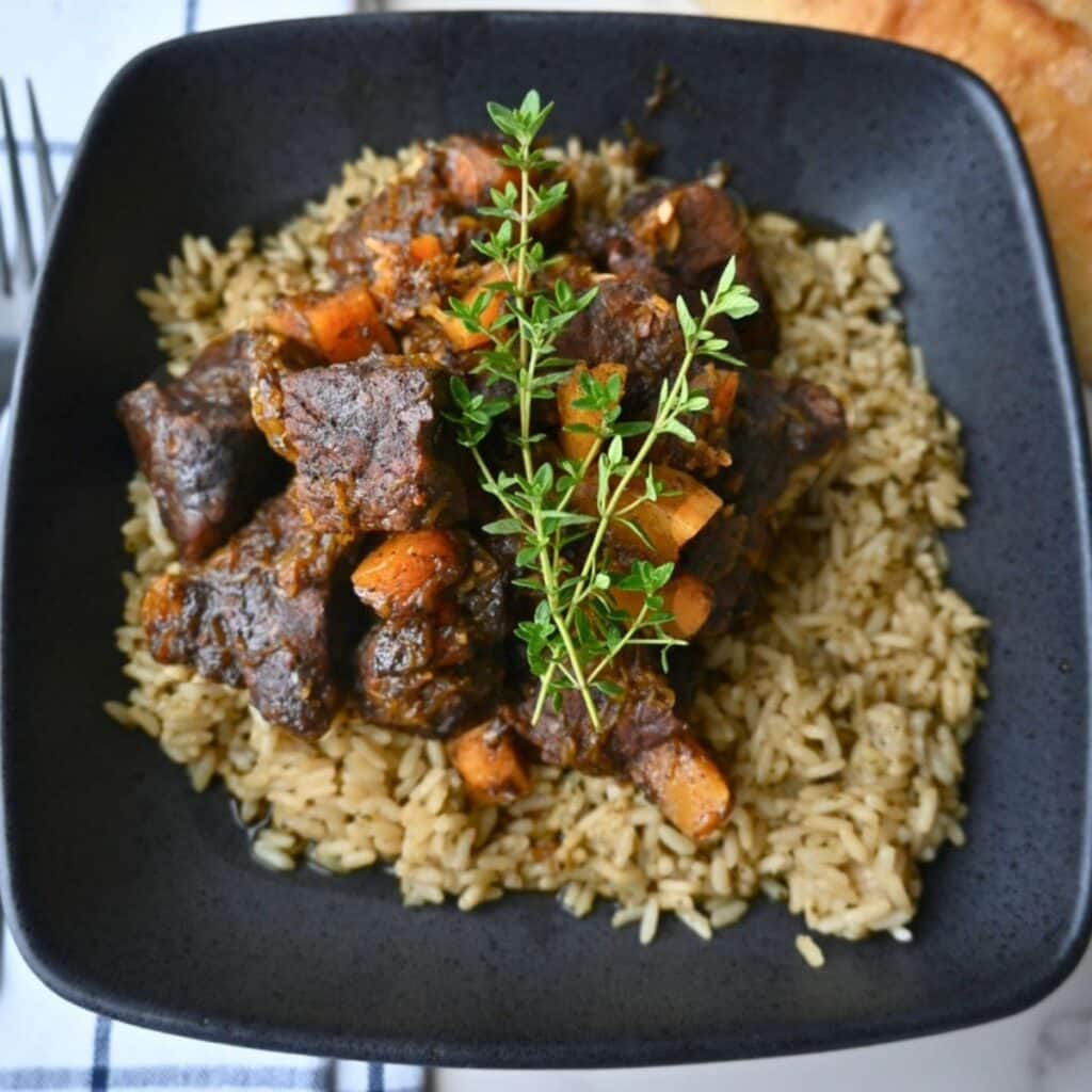 A black bowl holds seasoned rice topped with honey balsamic beef pieces and roasted butternut squash, finished with a sprinkle of fresh herbs.