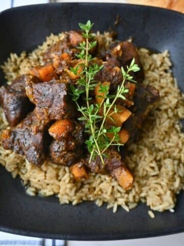 A black bowl holds seasoned rice topped with honey balsamic beef pieces and roasted butternut squash, finished with a sprinkle of fresh herbs.
