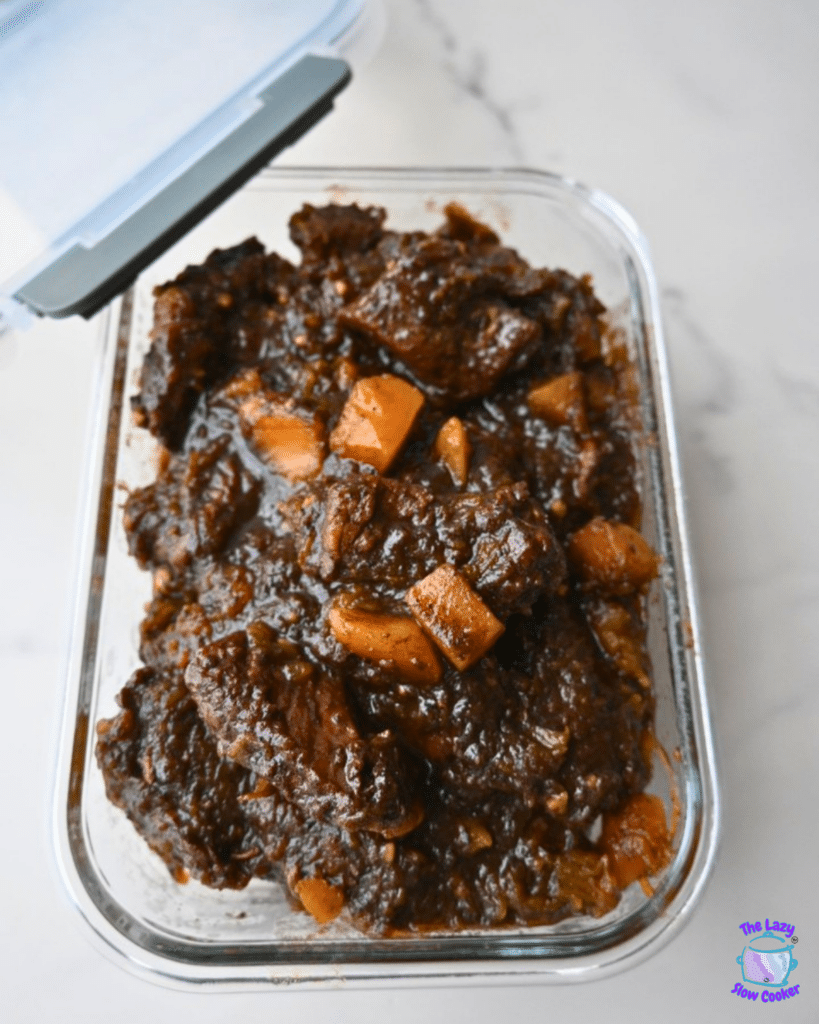 A clear glass container with the lid partly open holds honey balsamic beef and butternut squash stew, showing visible pieces of beef, orange carrots, and golden squash in a dark broth on a white surface.
