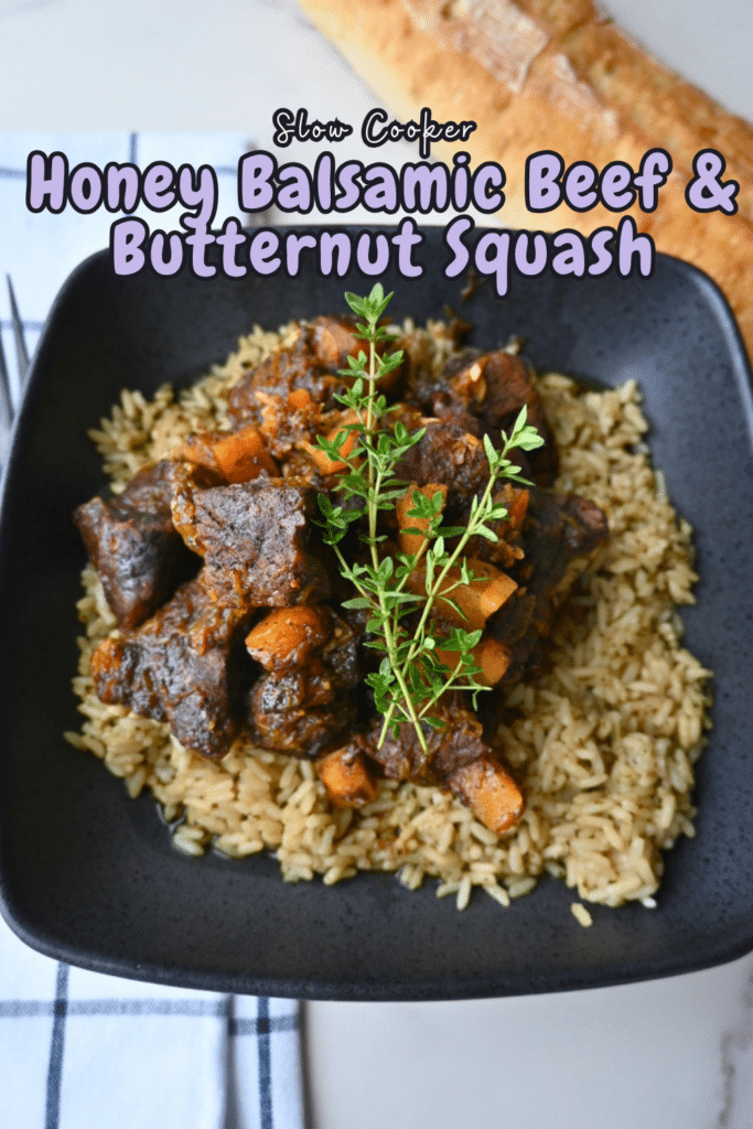 A black plate holds slow cooker honey balsamic beef, chunks of butternut squash, carrots, and a serving of white rice, all topped with fresh herbs. Above the plate is text that says “Slow Cooker Honey Balsamic Beef & Butternut Squash.” A sliced baguette sits in the background.