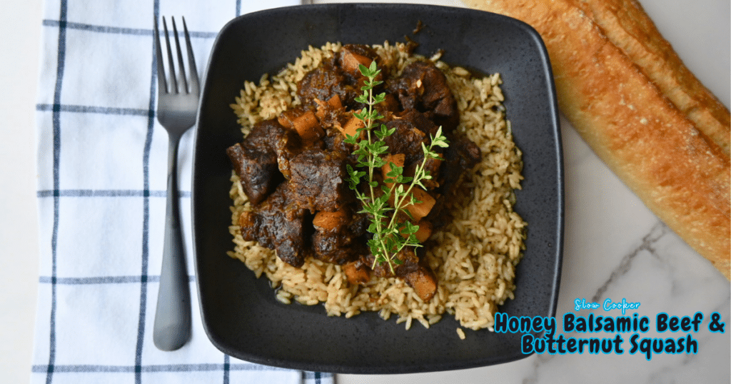 A square black bowl with honey balsamic beef and chunks of butternut squash served over rice, topped with fresh herbs. The bowl rests on a striped towel beside slices of bread. Blue text on the image says, “Slow Cooker Honey Balsamic Beef & Butternut Squash.”.