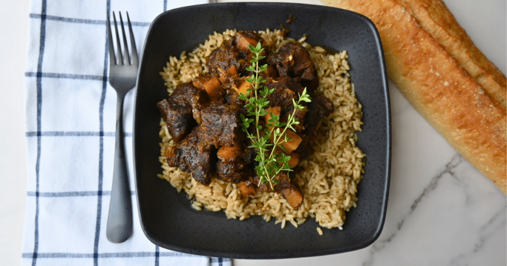 A black bowl filled with slow-cooked honey balsamic beef and butternut squash in a rich herb sauce sits atop a bed of rice. The bowl rests on a striped napkin, with a fork next to it and a piece of baguette on the side.