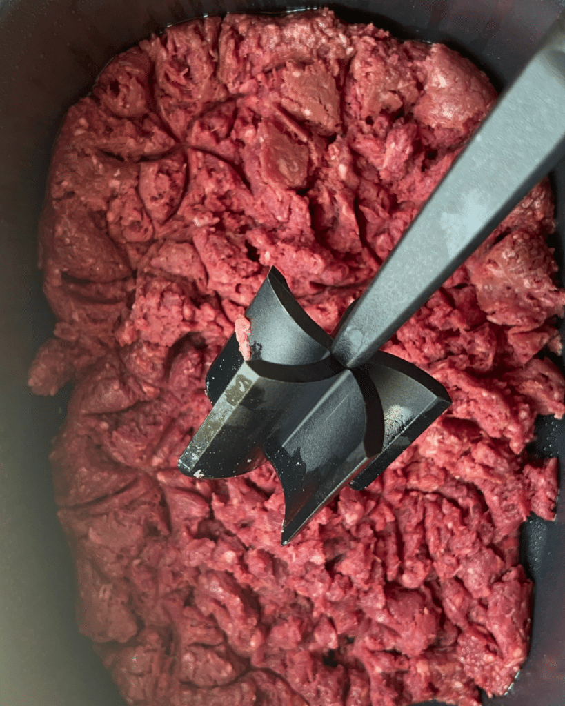 A black meat chopper presses down into raw ground beef in a dark container, starting to break up the meat for slow cooker American goulash.