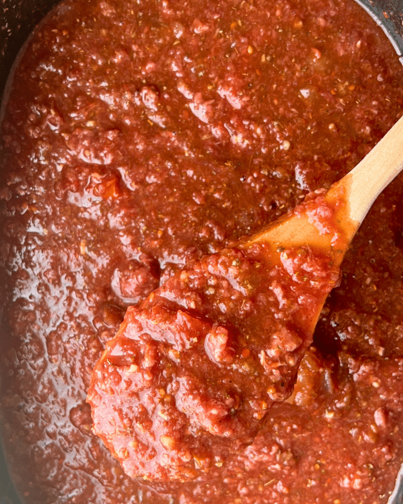 A wooden spoon scoops up thick, chunky tomato sauce with pieces of tomato, ground beef, and herbs from a pot, showing the rich texture typical of Slow Cooker American Goulash.