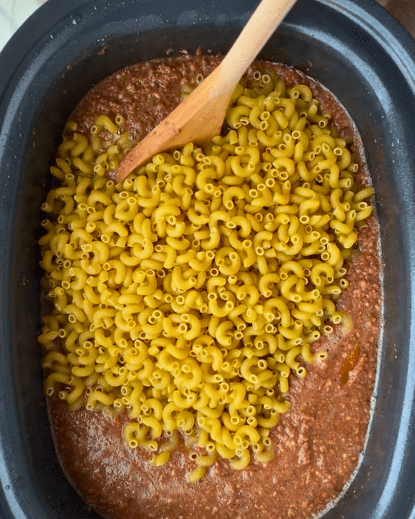 In a black slow cooker, ground beef and red sauce are layered at the bottom, with uncooked elbow macaroni spread on top. A wooden spoon rests in the mixture, about to be used for stirring this American goulash before cooking.