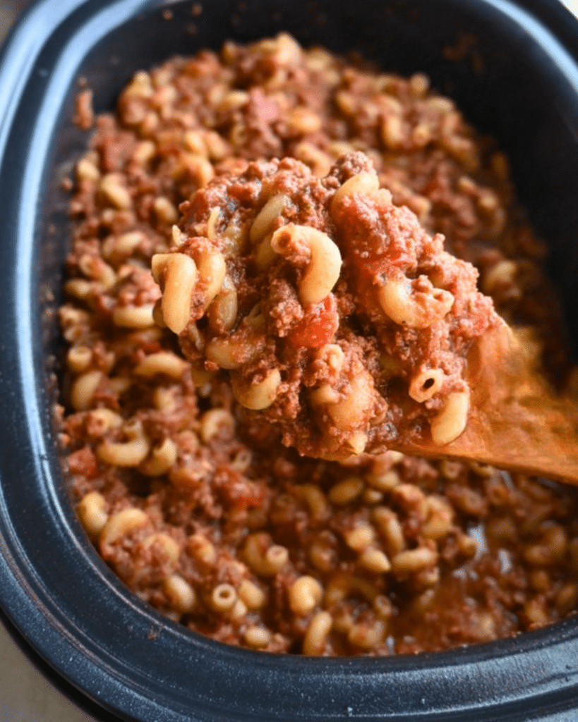 A wooden spoon lifts a serving of American goulash with macaroni, ground beef, and red tomato sauce from a black slow cooker.