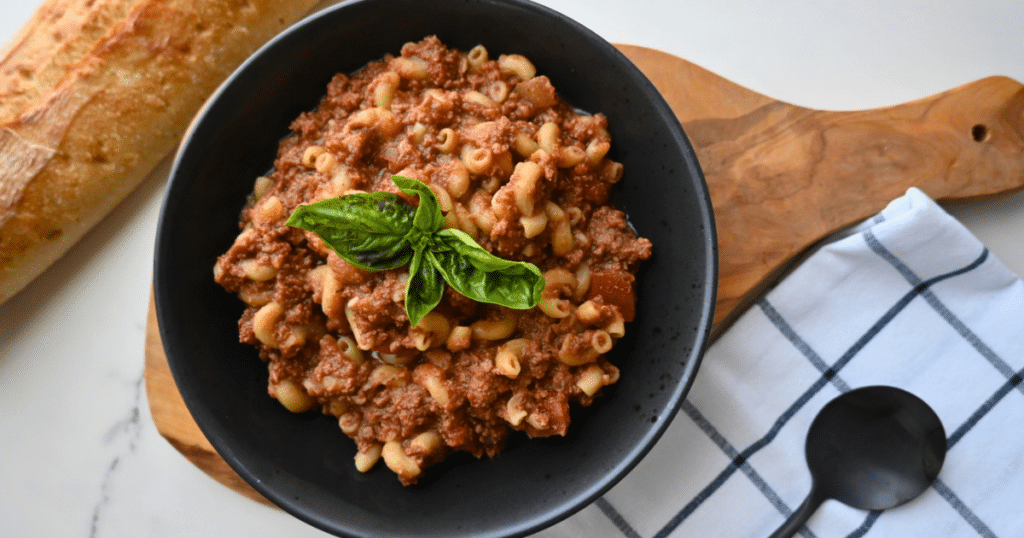 A black bowl holding macaroni pasta topped with Slow Cooker American Goulash and fresh basil is placed on a wooden board, with a loaf of bread, a spoon, and a checkered napkin beside it.
