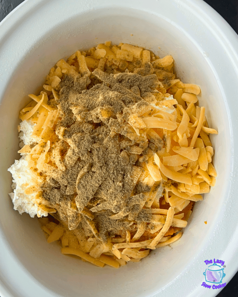 A white bowl holds shredded cheese, cream cheese, and assorted seasonings, all prepared to be mixed together for a slow cooker savory pumpkin dip. The Lazy Slow Cooker logo appears in the bottom right corner.