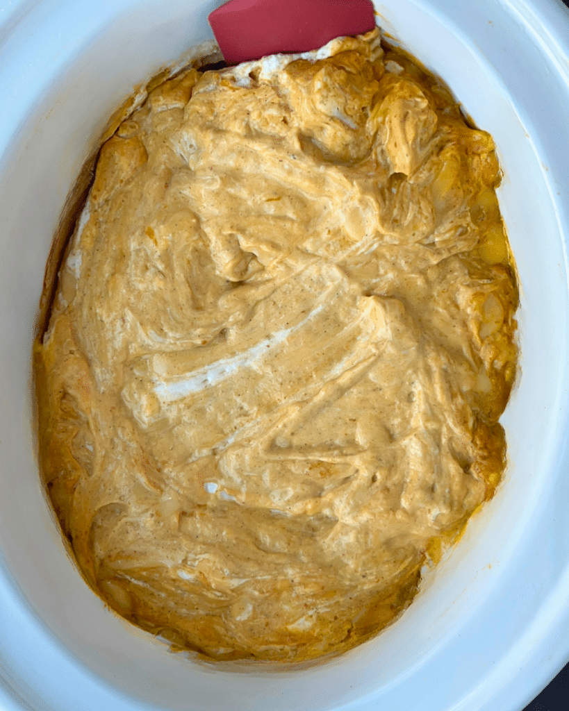 A white slow cooker holds a creamy, swirled pumpkin dip in light brown and white tones, with a red spatula resting at the edge.
