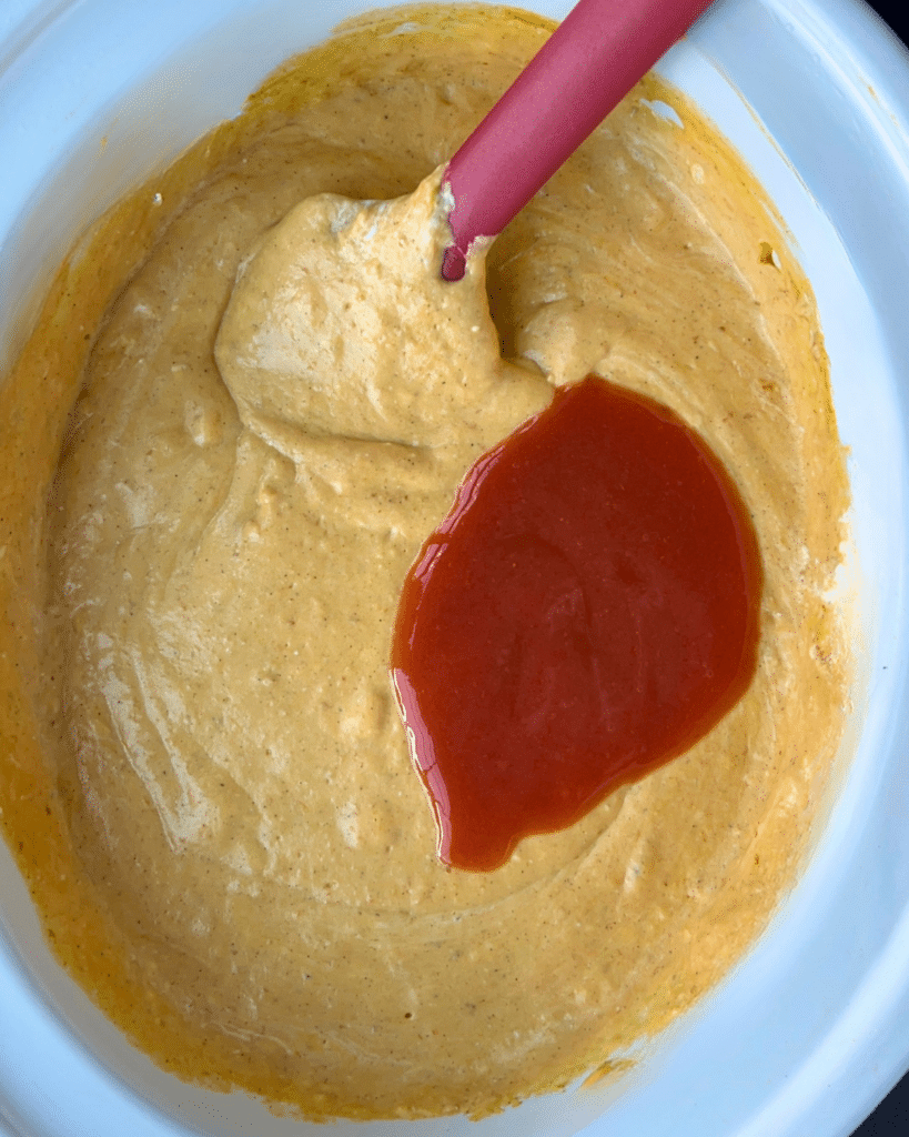 A mixing bowl holds thick yellow batter for savory pumpkin dip, with a red spatula resting inside and a layer of red sauce poured on top, not yet stirred in.