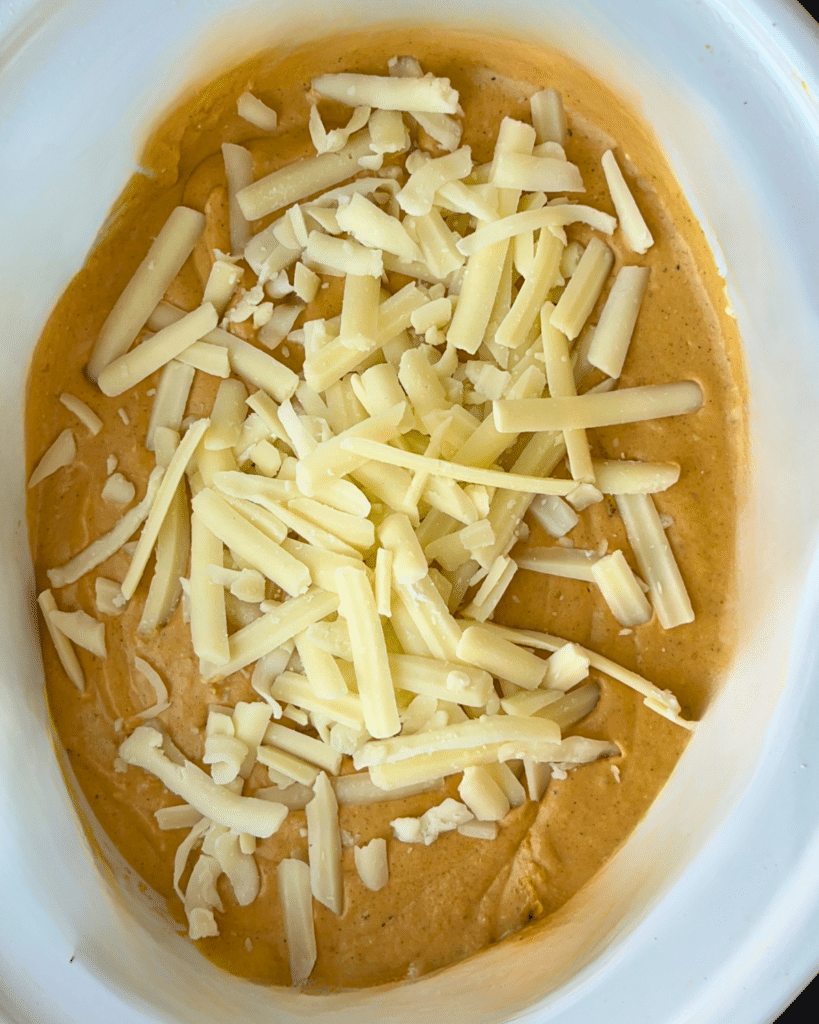 A white baking dish holds a smooth, orange mixture topped with shredded cheese, prepared to be combined or baked into a savory pumpkin dip using a slow cooker.