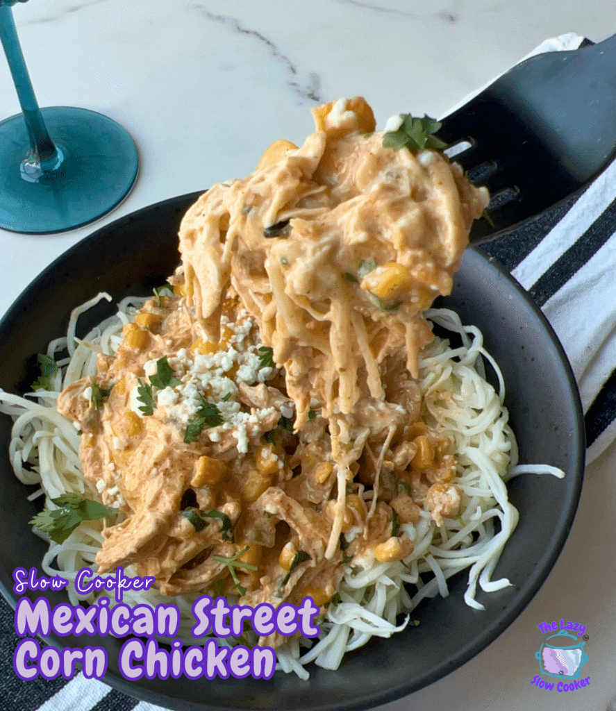 A black bowl filled with shredded chicken mixed with Mexican street corn is served over pasta and sprinkled with cheese and herbs. A fork lifts up some of the cheesy chicken and pasta. The words “Slow Cooker Mexican Street Corn Chicken” appear on the image.