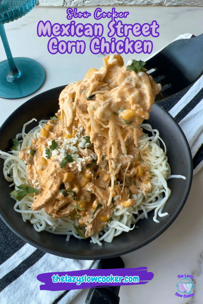 Shredded Mexican Street Corn Chicken, made in a slow cooker, is served over noodles in a black bowl and topped with cheese and cilantro. A fork lifts up some of the chicken and noodles. Text on the image advertises a slow cooker recipe from thelazyslowcooker.com.