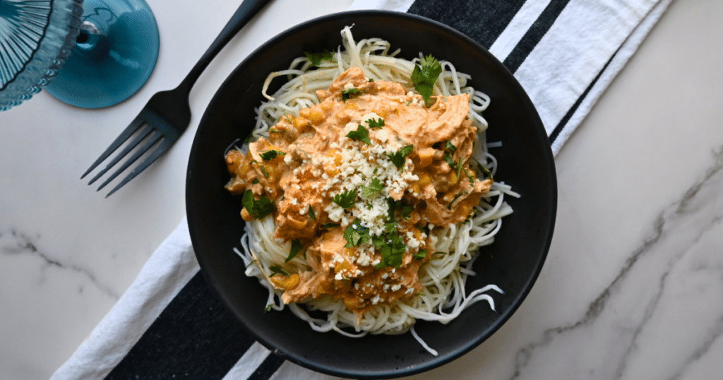A black bowl filled with spaghetti covered in creamy sauce, sprinkled with herbs and grated cheese, rests on a striped napkin beside a fork and blue glass on a marble table, evoking the look of Slow Cooker Mexican Street Corn Chicken pasta.