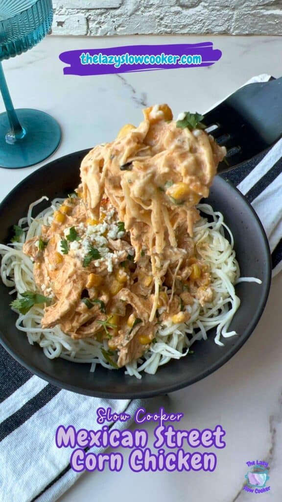 A black bowl holds shredded chicken in a creamy corn and cilantro sauce, served over pasta. A serving spoon is lifting some of the chicken mixture. The image includes text that says “Slow Cooker Mexican Street Corn Chicken.”.