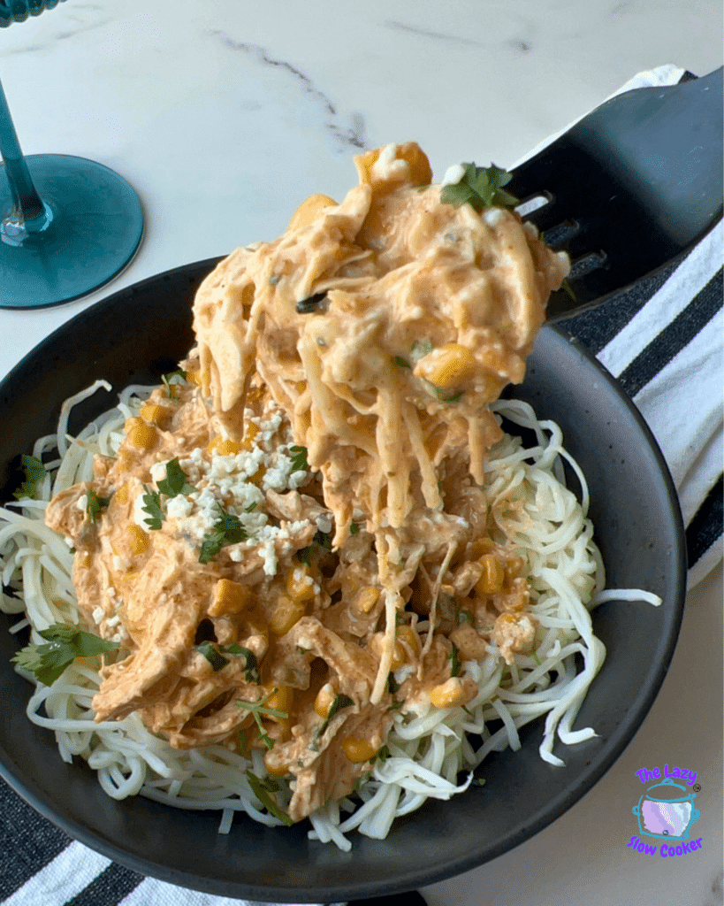 A fork lifts a serving of Slow Cooker Mexican Street Corn Chicken with creamy sauce, corn, and herbs over spaghetti noodles from a black bowl. In the background, there is a glass of wine and a striped napkin.