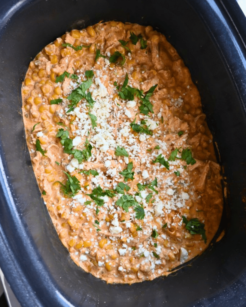 Tender chicken and sweet corn in a creamy sauce, topped with chopped cilantro and crumbled cheese, served inside a black slow cooker for Mexican Street Corn Chicken.
