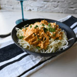 A black bowl holds Slow Cooker Mexican Street Corn Chicken, featuring shredded chicken in a creamy orange sauce over spaghetti noodles and topped with fresh herbs. The bowl sits on a striped towel next to a blue drinking glass.