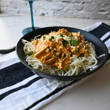 A black bowl holds Slow Cooker Mexican Street Corn Chicken, featuring shredded chicken in a creamy orange sauce over spaghetti noodles and topped with fresh herbs. The bowl sits on a striped towel next to a blue drinking glass.