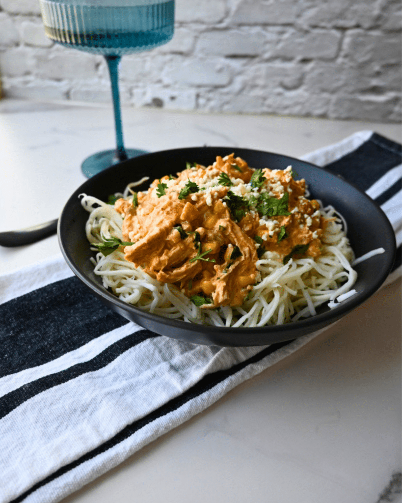 A black bowl holds Slow Cooker Mexican Street Corn Chicken, featuring shredded chicken in a creamy orange sauce over spaghetti noodles and topped with fresh herbs. The bowl sits on a striped towel next to a blue drinking glass.