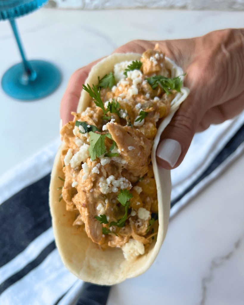 A hand holds a soft taco filled with creamy chicken, corn, crumbled cheese, and cilantro from a slow cooker Mexican Street Corn Chicken recipe. In the background, there is a blue glass and a striped napkin on a white surface.