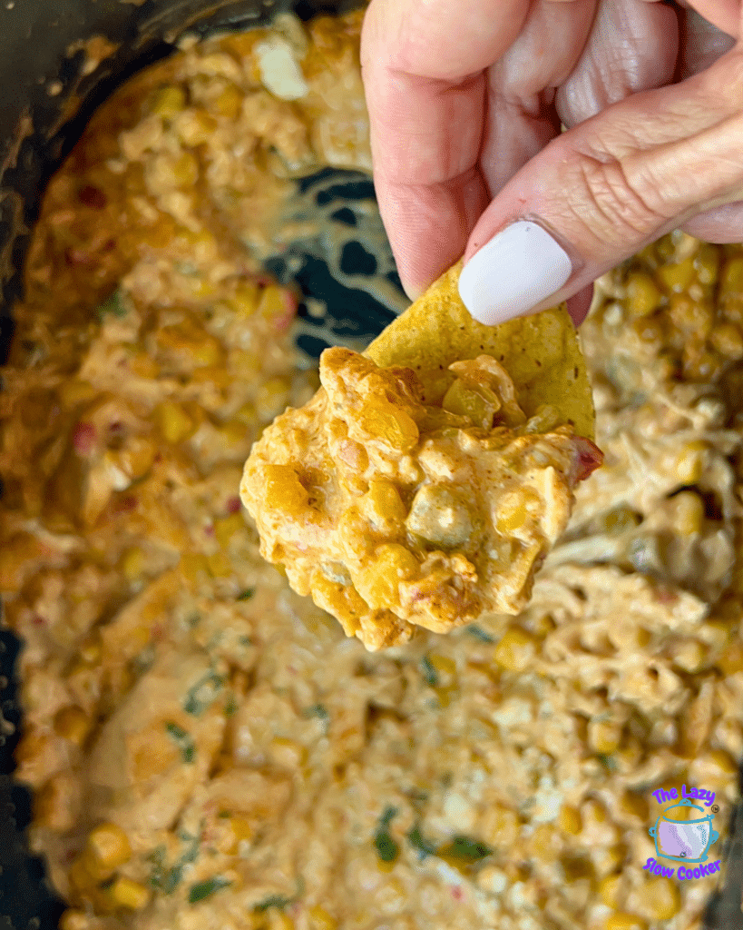 A hand with light pink painted nails lifts a tortilla chip covered in cheesy slow cooker Mexican Street Corn Chicken dip, showing corn, bell peppers, and herbs. Below, a bowl is filled with more of the same creamy dip.