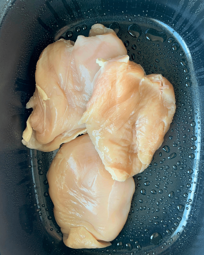 Three raw, boneless, skinless chicken breasts sit inside a black slow cooker pot with visible water droplets on the sides, ready to be cooked for Mexican Street Corn Chicken.