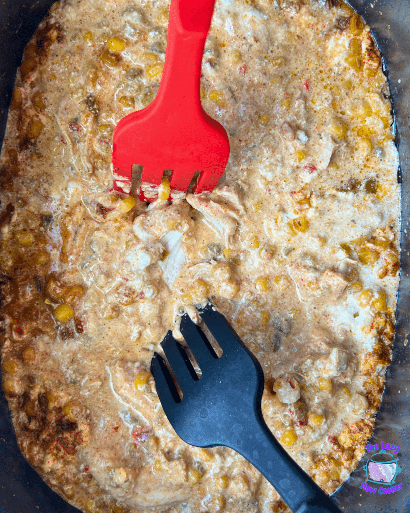 A large pan holds a creamy mixture of corn, shredded chicken, and spices being stirred with red and black utensils. This visually resembles Slow Cooker Mexican Street Corn Chicken.