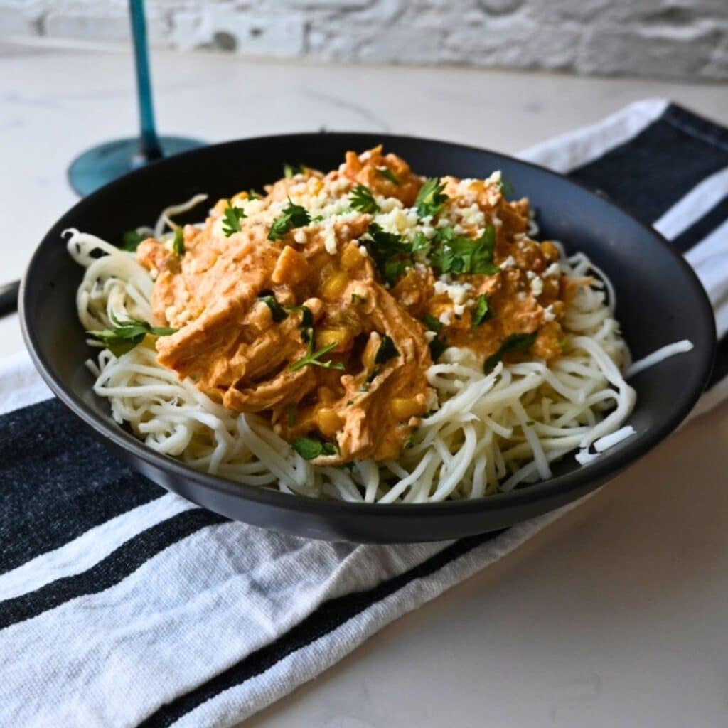 A black bowl filled with creamy Mexican Street Corn Chicken made in a slow cooker, featuring shredded chicken over spaghetti noodles and topped with chopped herbs and grated cheese, is placed on a striped napkin on a white tabletop.