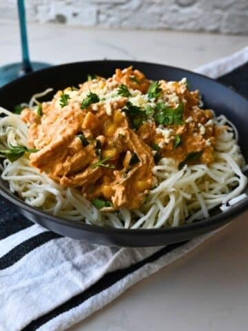 A black bowl filled with creamy Mexican Street Corn Chicken made in a slow cooker, featuring shredded chicken over spaghetti noodles and topped with chopped herbs and grated cheese, is placed on a striped napkin on a white tabletop.