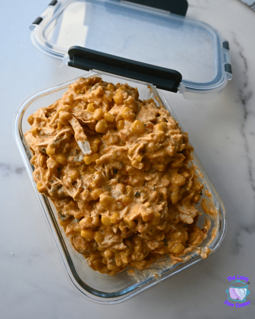 A glass dish on a white countertop holds Slow Cooker Mexican Street Corn Chicken, showing a thick, creamy blend of corn, shredded chicken, and visible spices mixed together.