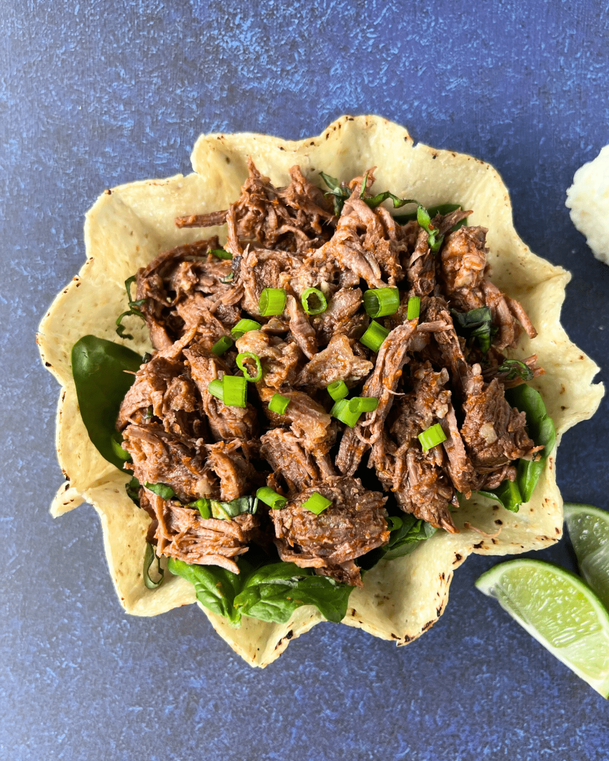 A tortilla bowl filled with slow-cooked shredded beef sits on a bed of fresh spinach, topped with chopped green onions. Lime wedges are arranged beside the bowl, all set on a blue textured surface.