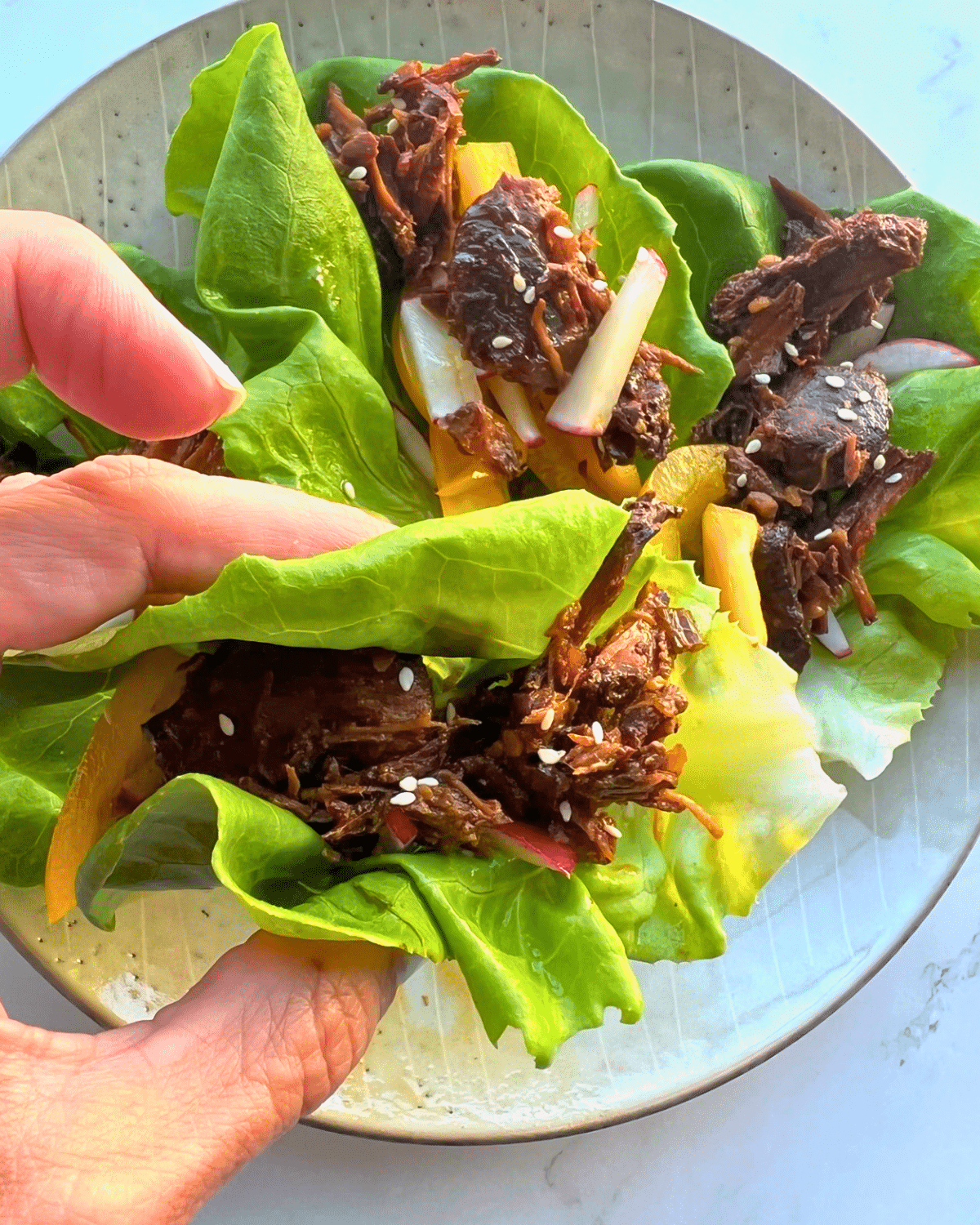 A hand holds a lettuce wrap filled with shredded meat, sliced radish, yellow bell pepper strips, and sesame seeds. In the background, several more slow cooker lettuce wraps with the same colorful fillings are arranged on a plate.