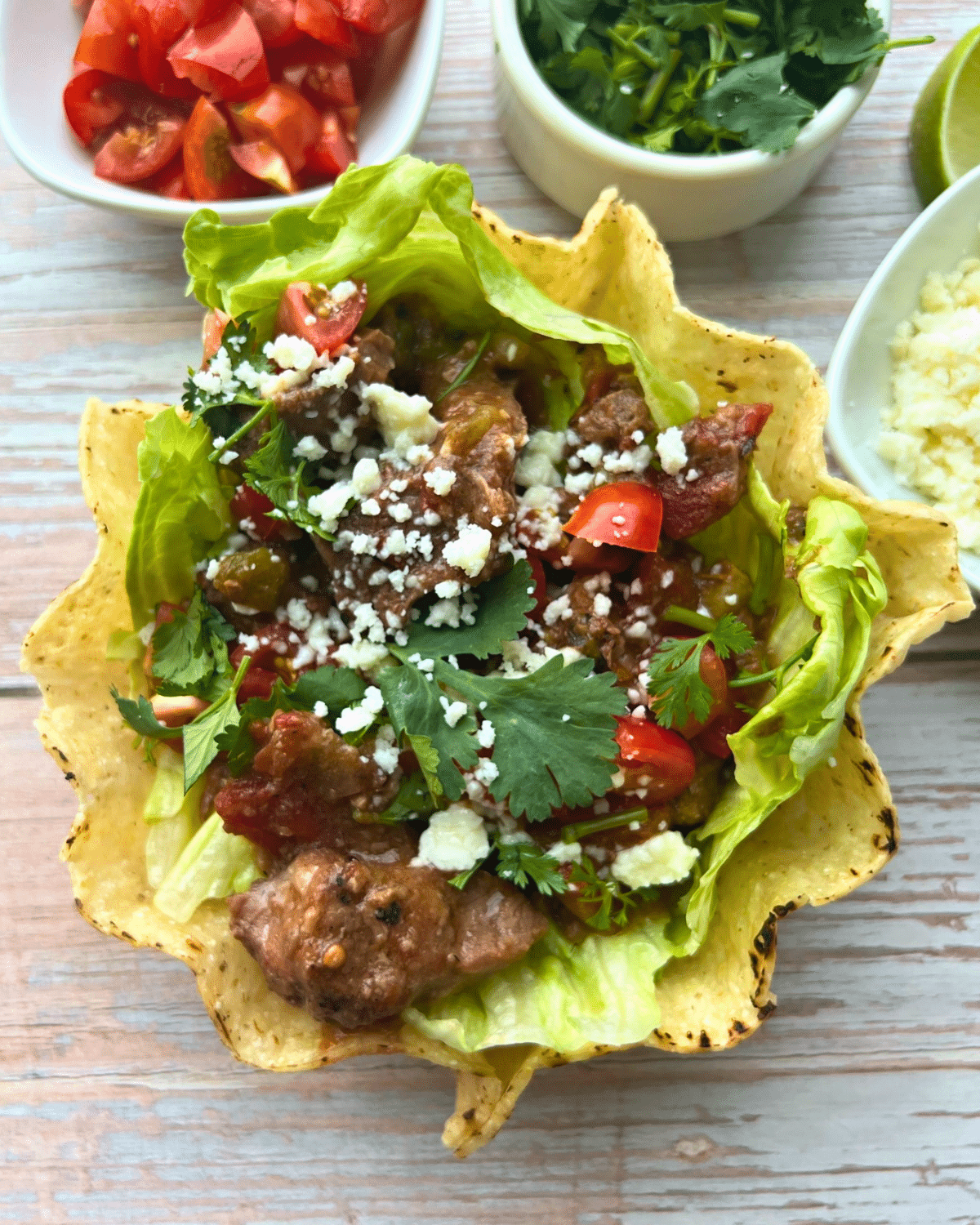 Carne picada in a crisp tortilla bowl. Crisp layers of lettuce, slow cooked pieces pieces of beef, diced tomatoes, fresh cilantro, and crumbled cheese. Small bowls of extra toppings sit beside the main dish.
