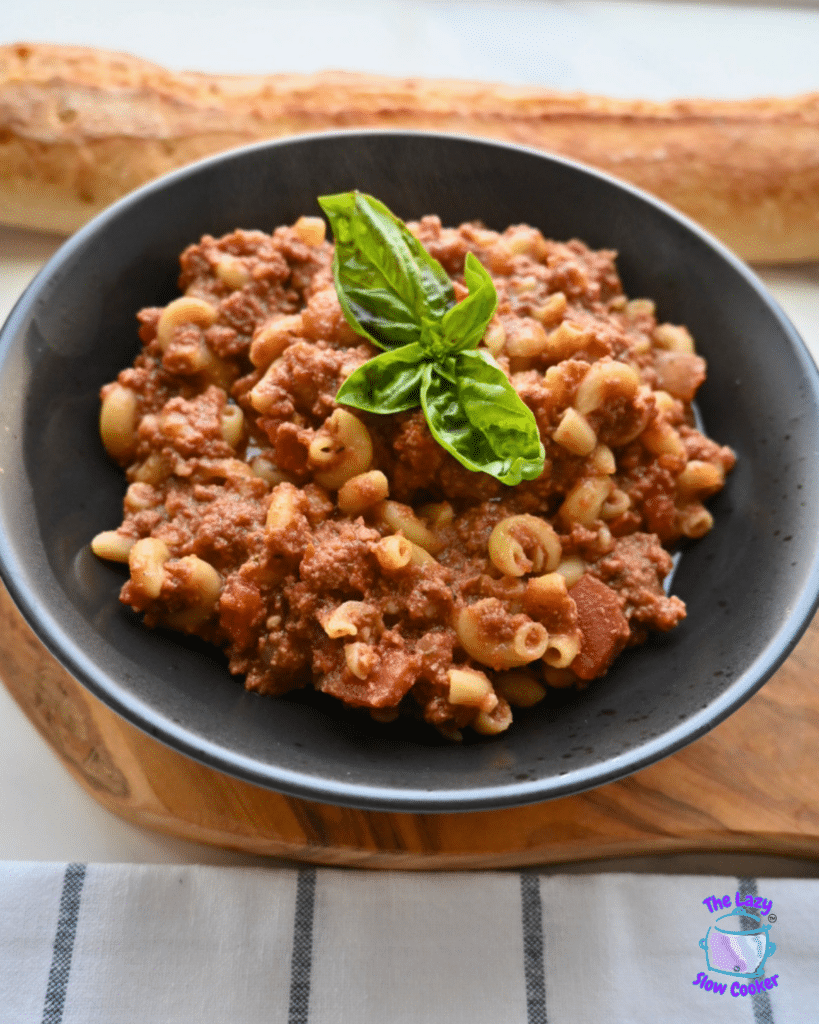 A black bowl of macaroni beef chili, made in a slow cooker and garnished with fresh basil, sits on a wooden board. In the background, there is a baguette and a striped cloth.
