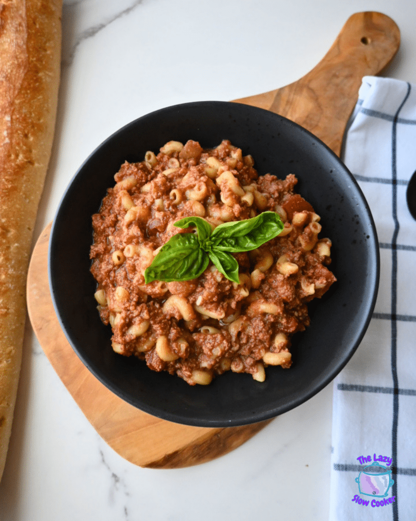 A black bowl filled with macaroni and meat sauce, topped with fresh basil, sits on a wooden board beside a loaf of bread and a striped napkin. The pasta dish resembles slow cooker American goulash.