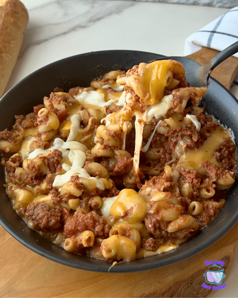 A spoon lifts a serving of cheesy beef and macaroni casserole from a skillet, with melted cheese stretching between the spoon and the dish. In the background, there is a loaf of bread and a kitchen towel.