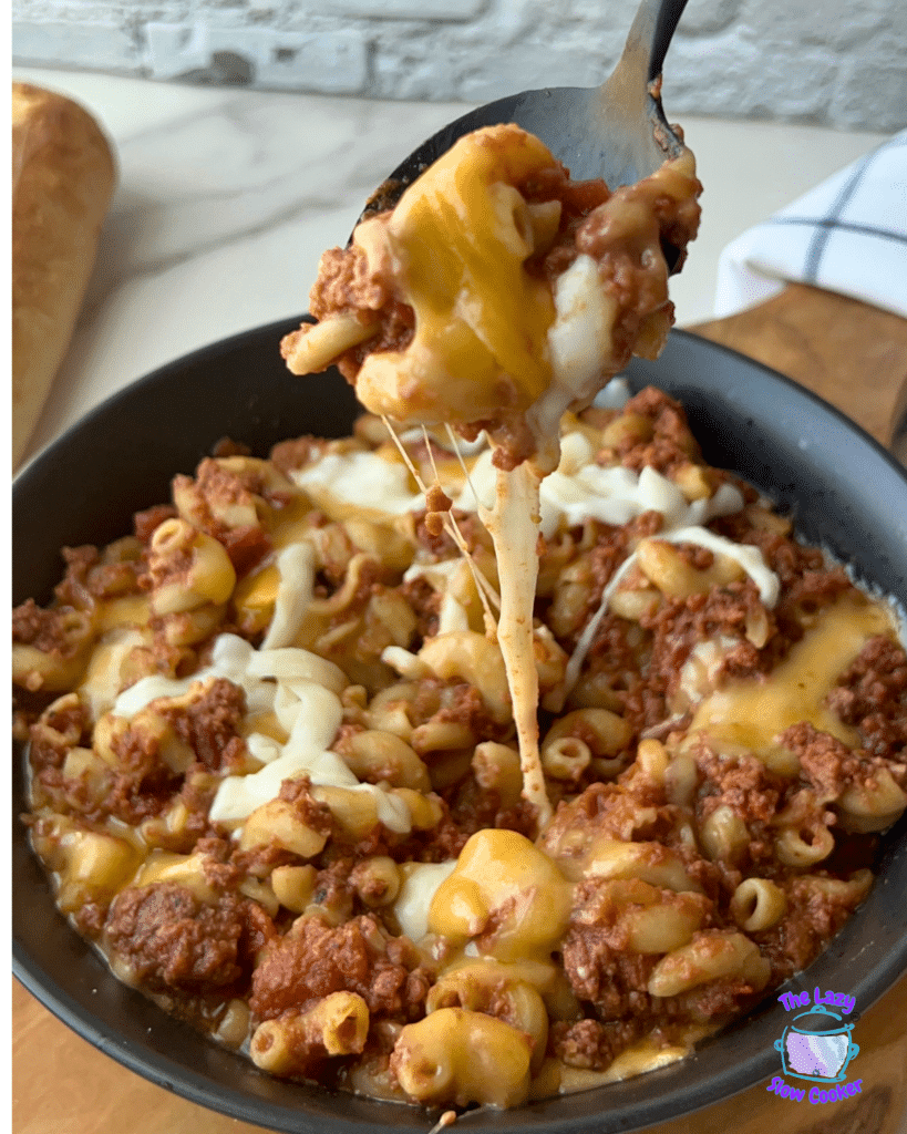 A close-up shows Slow Cooker American Goulash being scooped from a bowl, with melted cheese stretching between the spoon and the pasta. Ground beef, elbow macaroni, and gooey cheddar and mozzarella are visible in the scoop. A loaf of bread sits blurred in the background.